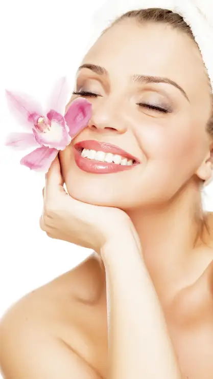 Mujer sonriendo sujetando una bonita flor rosa cerca de su cara representando que tiene una buena higiene facial